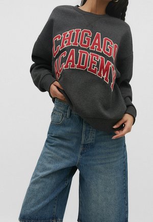 Personne portant un sweat-shirt gris foncé avec "CHICAGO ACADEMY" en grandes lettres rouges et un short en jean bleu ample, debout sur fond blanc.