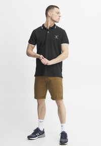 Černé polo tričko s bílým límcem a akcenty na rukávech, logo na hrudi, v kombinaci s khaki zkrácenými šortkami a námořnickými teniskami. Bílé ponožky jsou viditelné.