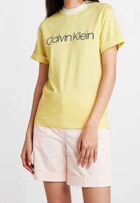 Žluté pruhované tričko s kulatým výst�řihem a krátkými rukávy, s černým logem "Calvin Klein". Nosené s světle růžovými šortkami.