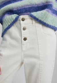 Jeans de tiro alto color crema con tres botones de metal que presentan un diseño de calavera. Llevados bajo un suéter de punto texturizado y multicolor.