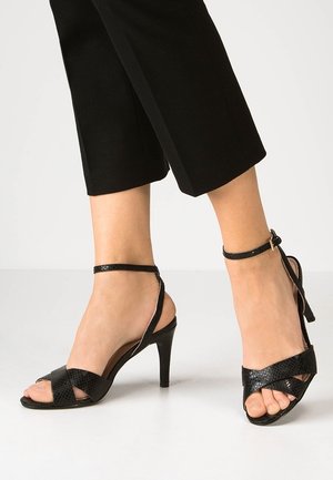 Voeten met zwarte sandalen met hoge hak, enkelbandjes en gekruiste voorbandjes, gecombineerd met zwarte cropped broeken.