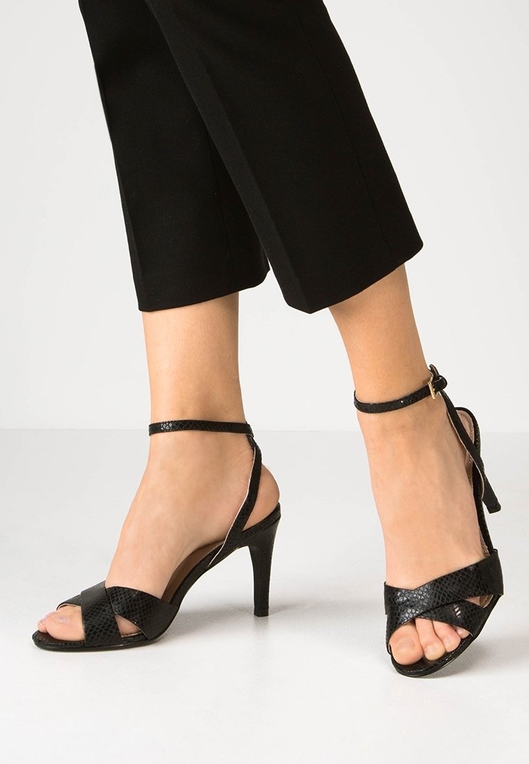 Pieds portant des sandales noires à talons hauts avec des brides à la cheville et des brides croisées à l'avant, associés à un pantalon noir cropped.