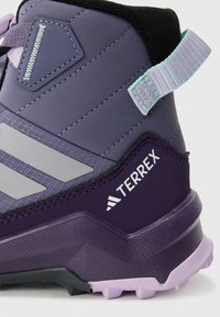 Botte de randonnée violette avec une tige texturée, des accents argentés et une semelle en caoutchouc. Présente un tirant à l'arrière et un logo "TERREX" sur le côté.