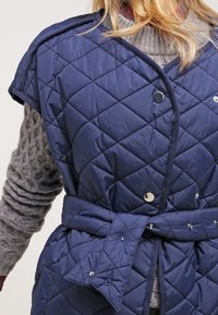 Marinblå vadderad väst med krage, tryckknappar och bälte. Har diamantmönster och mjuk textur, lagd över en grå stickad tröja.