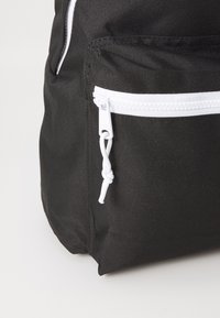 Mochila preta com fecho branco, feita de tecido durável. Apresenta um bolso frontal com uma alça de puxar para fácil acesso e uma superfície texturizada.
