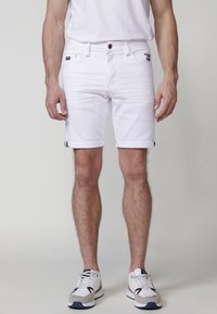 Pantalones cortos de mezclilla blanca con puños enrollados, que presentan un cierre frontal con botón, dos bolsillos laterales y un pequeño parche con el logo en la cadera izquierda.