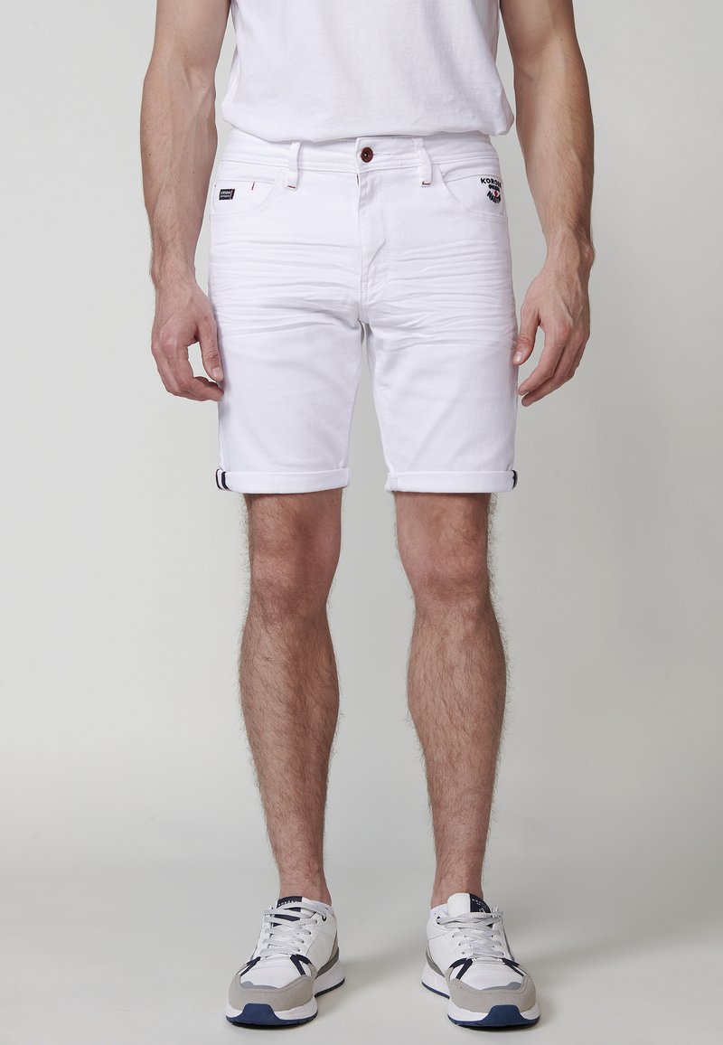 Pantalones cortos de mezclilla blanca con puños enrollados, que presentan un cierre frontal con botón, dos bolsillos laterales y un pequeño parche con el logo en la cadera izquierda.