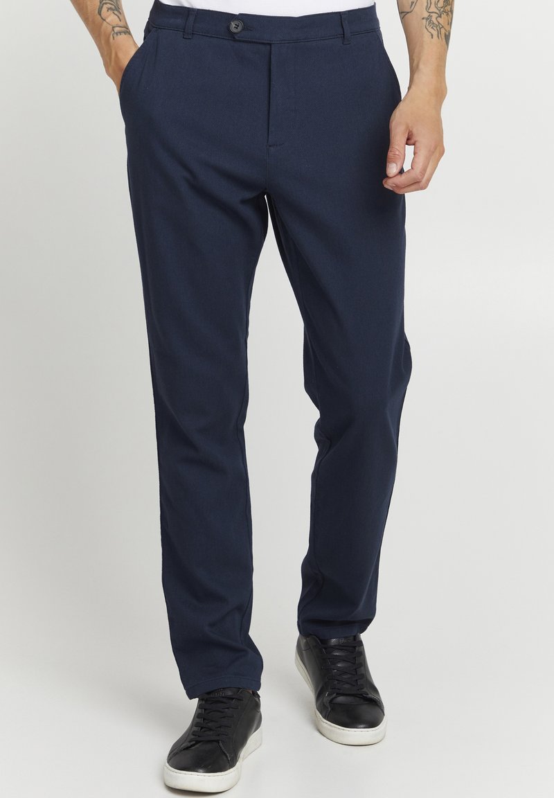 Homme portant un pantalon bleu marine et des baskets noires, debout les mains détendues le long du corps, sur un fond uni.