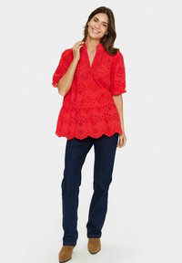 Rdeča bluzka z očesci, kratkimi napihnjenimi rokavi, volanastim robom in V-izrezom, kombinirana z temno modrimi jeans hlačami in rjavimi gležnjarji.