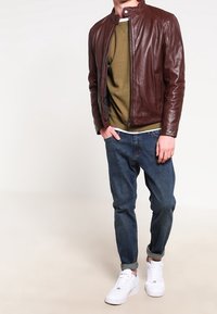 Brun læderjakke med ståkrave, olivengrøn T-shirt, blå jeans og hvide sneakers. Jakken har et slankt design og en frontlynlås.