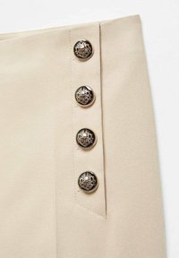 Jupe en tissu beige ornée de quatre boutons décoratifs noirs et dorés sur le côté, avec une texture lisse et un design ajusté.