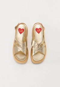 Sandálias plataforma com tiras cruzadas em dourado e sola texturizada. Logótipo em forma de coração "LOVE MOSCHINO" em vermelho na palmilha. Fivela ajustável no tornozelo.