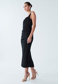 Vestido negro sin mangas con detalle fruncido y diseño ajustado, combinado con sandalias de tacón alto doradas con tiras, en una modelo sobre un fondo claro.