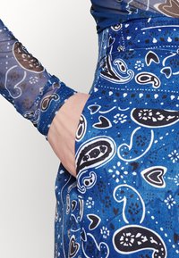 Detailní záběr na ruku v kapse modré sukně s černobílými vzory paisley a srdcí, doplněné odpovídajícím dlouhým rukávem.