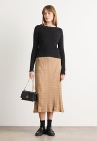 Černý pletený svetr, dlouhá hnědá skládaná sukně, černá crossbody kabelka, černé mokasíny a kotníčkové ponožky na světlém pozadí.