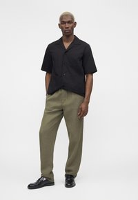 Homme aux cheveux blonds courts portant une chemise noire à manches courtes boutonnée, un pantalon vert olive et des chaussures en cuir noir, debout sur un fond blanc.