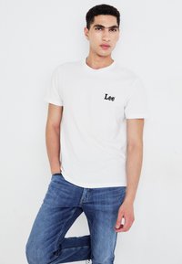 Camiseta blanca de algodón con un logo negro de "Lee" en el lado izquierdo del pecho, combinada con unos jeans de mezclilla azules. Cuello redondo sencillo y mangas cortas.