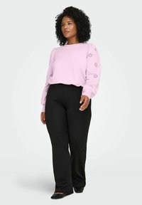 Svetlo roza pulover z okroglimi detajli na rokavih, kombiniran s črnimi, oprijetimi hlačami in ravnimi črnimi čevlji. Gladek material, sproščen kroj.