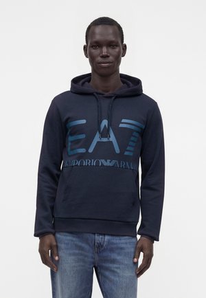 Mężczyzna w granatowej bluzie z kapturem Emporio Armani z logo EA7 i jasnoniebieskich dżinsach, stojący na jednolitym białym tle.