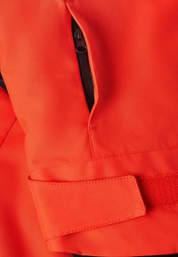 Veste orange vif en tissu durable avec une poche zippée et un détail de sangle en velcro. Texture lisse, design structuré.