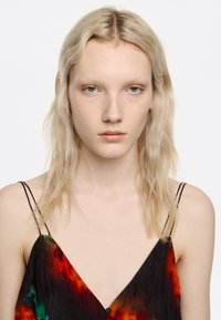 Junge blasse Frau mit langen blonden Haaren trägt ein ärmelloses schwarzes Kleid mit roten und grünen Mustern und blickt direkt in die Kamera.