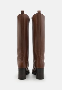 Bottes en cuir marron jusqu'au genou avec une finition lisse, fermeture éclair et un talon empilé. Présente des coutures verticales sur les côtés.