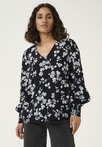Blusa floreale con scollatura a V, maniche lunghe a palloncino e polsini arricciati. Sfondo blu scuro con motivo floreale bianco; tessuto dalla consistenza morbida.