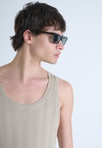 Top a fascia beige con righe e scollatura ampia. Occhiali da sole neri con montatura angolare. Carnagione chiara e capelli scuri di lunghezza media. Sfondo neutro.