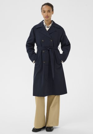 Frau trägt einen marineblauen Trenchcoat mit Gürtel über einem karierten Hemd und beigen Hosen mit weitem Bein, steht mit den Händen in den Taschen des Mantels.