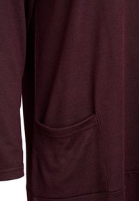 Primo piano di una manica e della parte inferiore frontale di un capo in maglia bordeaux scuro con una piccola tasca rettangolare.