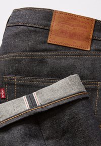 Dark denim jeans featuring a leather patch with embossed details, orange stitching, and contrasting gray lining with red accents.