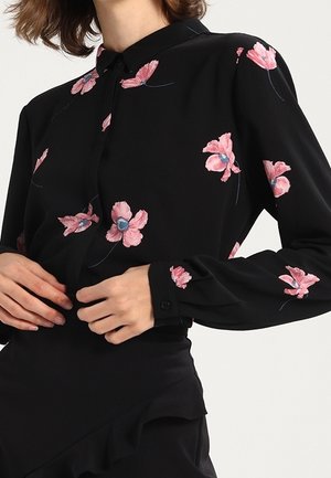 Kvinde iført en sort bluse med pink blomsterbroderi og en sort nederdel med flæsedetalje, hænder justerer blusen.