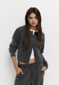 Cardigan gri din lână cu nasturi în față și mansete cu aspect texturat, purtat peste o cămașă albă, asortat cu pantaloni gri lejerați cu şnur.