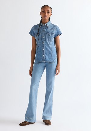 Femme aux cheveux tressés debout, portant une chemise en jean à manches courtes, un jean évasé bleu clair et des mocassins marron sur fond blanc.