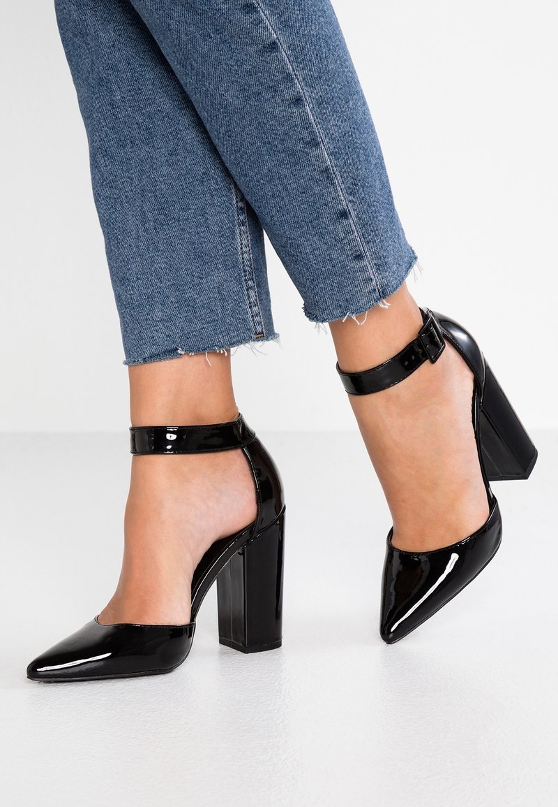 Des chaussures à talons hauts en cuir verni noir avec un bout pointu et une bride à la cheville, présentant un large talon. Portées avec un pantalon en denim bleu court.