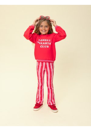 Meisje draagt een rood sweatshirt met de tekst "Lonely Hearts Club", rood gestreepte broek, rode sneakers, en houdt rode zonnebril boven haar hoofd.