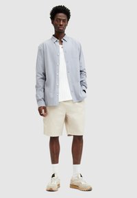 Lichtblauw overhemd met lange mouwen, witte textuur t-shirt, beige shorts en beige sneakers met een rubberen zool. Kort, krullend haar.