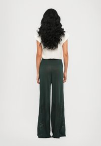 Zelene široke hlače z ribasto elastiko in prozornimi detajli na robu. Material se zdi lahek in teksturiran. Prikazana je zadnja stran.