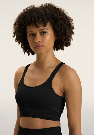Femme aux cheveux bouclés portant un soutien-gorge de sport noir, debout devant un fond clair uni, regardant la caméra avec une expression neutre.