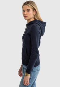 Jauna sieviete tērpusies tumši zilā rāvējslēdzēja džemperī un gaismazilās džinsu biksēs, stāvot sāniski pret vienkāršu gaišu fonu.