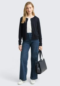 Marineblauwe cardigan, witte top en wijde blauwe jeans; gecombineerd met witte sneakers en een zwarte tas. Glad materiaal met knopen op de cardigan.