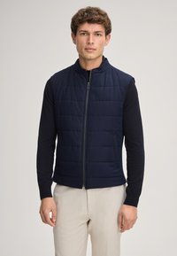 Granatowa pikowana kamizelka z wysokim kołnierzem, zamkiem błyskawicznym z przodu i bocznymi kieszeniami. Nosi się ją na czarnej koszuli z długim rękawem, zestawiona z jasnymi spodniami.