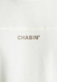 Balta medvilninė marškinėliai su tekstūruotu, šviesiai pilku "CHASIN'" logotipu. Audinys yra švelnus ir lygus, su tvarkingais siūlių detalėmis.