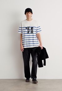 Weißes kurzärmliges T-Shirt mit blauen horizontalen Streifen, versehen mit einem schwarzen Grafikdesign. Kombiniert mit schwarzen Jeans und einer schwarzen Jacke steht der Träger vor einem schlichten Hintergrund.