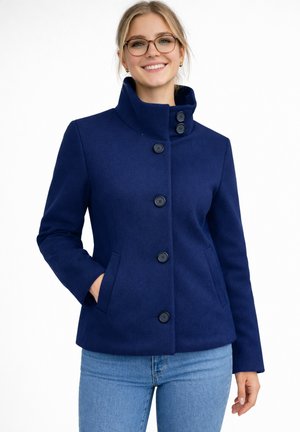 Lachende vrouw met een bril en een donkerblauwe jas met knopen en een hoge kraag, handen in de zakken, gecombineerd met lichtblauwe jeans.