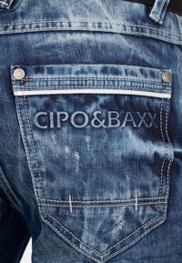 Jeans en denim bleu avec un logo sur la poche arrière, des coutures contrastées, un léger délavage et des détails de quincaillerie décoratifs.