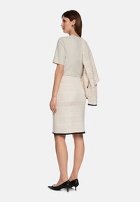 Helle beige Bluse mit kurzen Ärmeln, kombiniert mit einem strukturierten cremefarbenen Rock mit aufwendigen Mustern und schwarzem Saum. Schwarze spitze Absätze runden das Outfit ab.