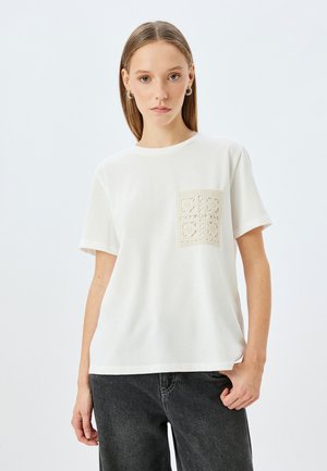 T-shirt blanc à manches courtes avec un détail de poche en dentelle beige texturée, dotée d'un col rond et d'une coupe décontractée.