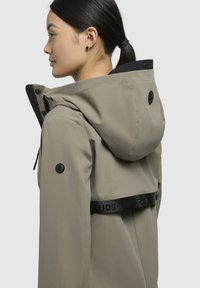 Giacca impermeabile color beige chiaro con cappuccio, cintura nera regolabile, dettagli con logo e bottoni neri visibili sulle maniche e sul retro.