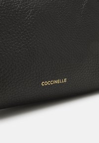 Surface en cuir texturé noir avec un petit nom de marque métallique doré "COCCINELLE" embossé près du bord inférieur.